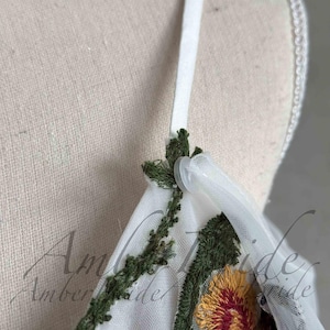 May include: Close-up of a white tulle fabric with green and yellow floral embroidery. The fabric has a white satin strap with a plastic button.