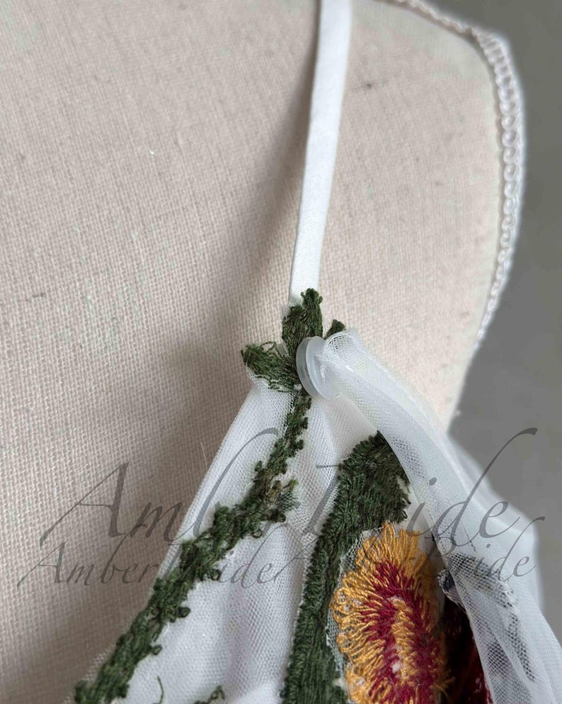 May include: Close-up of a white tulle fabric with green and yellow floral embroidery. The fabric has a white satin strap with a plastic button.