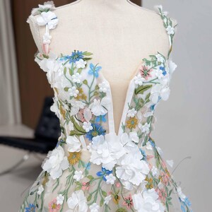 May include: A white tulle dress with a plunging neckline and floral embroidery. The dress features 3D flowers in white, blue, pink, and yellow.
