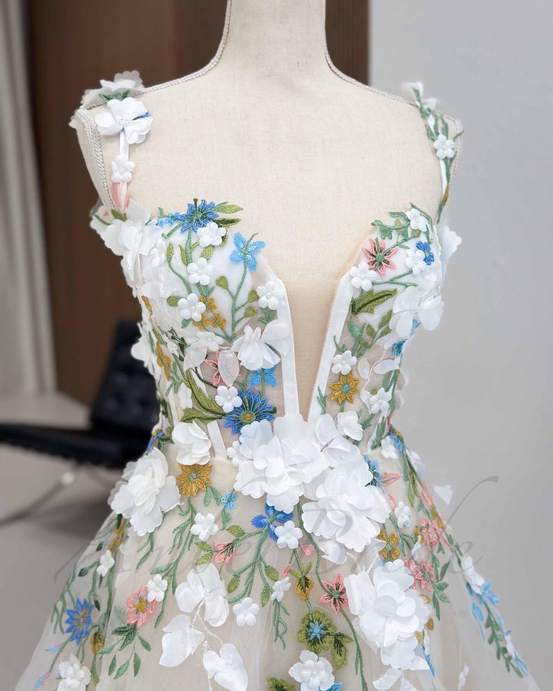 May include: A white tulle dress with a plunging neckline and floral embroidery. The dress features 3D flowers in white, blue, pink, and yellow.