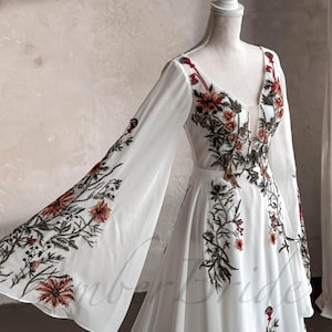 May include: A white maxi dress with long, flowing sleeves and a V-neckline. The dress is adorned with floral embroidery in shades of red, orange, and green.