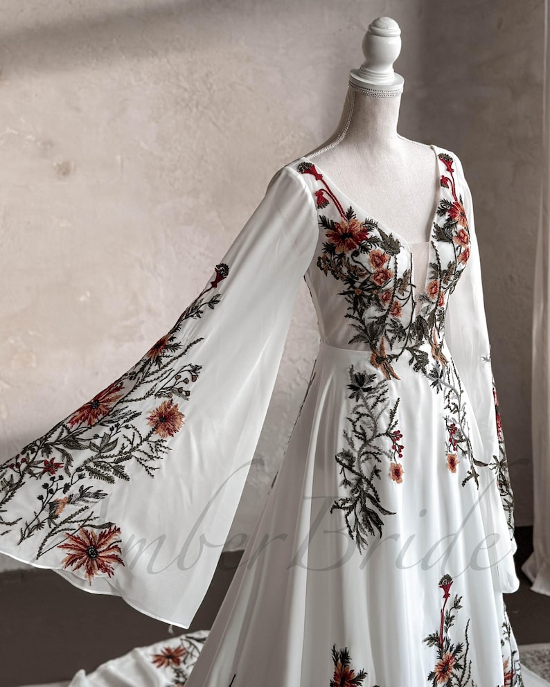 May include: A white maxi dress with long, flowing sleeves and a V-neckline. The dress is adorned with floral embroidery in shades of red, orange, and green.