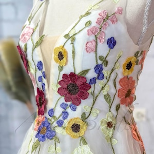 May include: A white tulle dress with colorful embroidered flowers. The flowers are in shades of red, yellow, blue, pink, and orange. The dress has a V-neckline and sleeveless design.