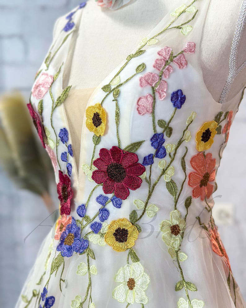 May include: A white tulle dress with colorful embroidered flowers. The flowers are in shades of red, yellow, blue, pink, and orange. The dress has a V-neckline and sleeveless design.
