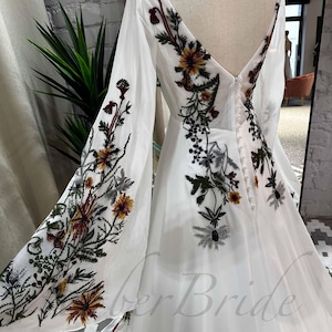 May include: A white wedding dress with long sleeves and a V-neckline. The dress is adorned with floral embroidery in shades of green, brown, and yellow.