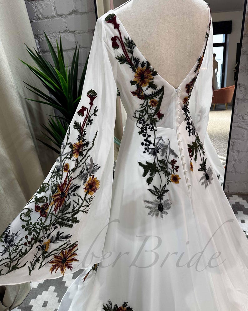 May include: A white wedding dress with long sleeves and a V-neckline. The dress is adorned with floral embroidery in shades of green, brown, and yellow.