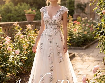 Puede incluir: Un vestido de novia color crema con bordado floral. El vestido presenta un escote en V, mangas cortas y una falda fluida. El bordado incluye patrones florales rosas y verdes. El vestido se usa al aire libre en un jardín con el texto "Amber Bride".