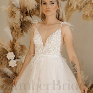 May include: A white bridal gown with a plunging neckline, delicate lace detailing, and a feather trim. The gown has a flowing tulle skirt and a fitted bodice.