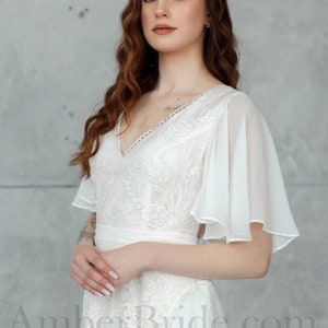 May include: A white lace wedding dress with a V-neckline and sheer, flowing sleeves. The dress has a fitted bodice and a flowing skirt. The model is wearing the dress with her hair in loose waves.