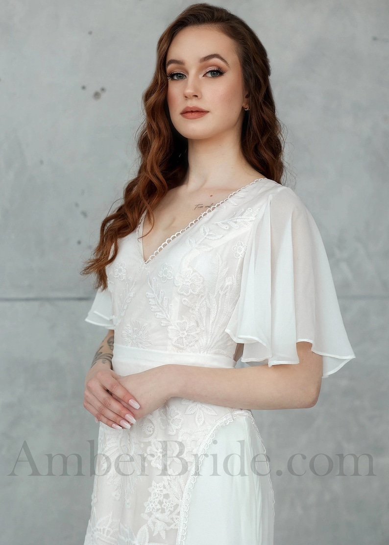 May include: A white lace wedding dress with a V-neckline and sheer, flowing sleeves. The dress has a fitted bodice and a flowing skirt. The model is wearing the dress with her hair in loose waves.