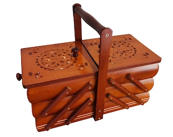 May include: A wooden sewing box with a tiered, expandable design and a central carrying handle. The top lids feature carved floral patterns, and the structure includes multiple compartments connected by hinged wooden arms in a warm brown finish.