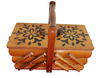 May include: A wooden sewing box with a tiered accordion-style design and a central carrying handle. The top lids feature dark, intricate floral carvings on a light brown wood surface, complemented by small round knobs for opening the compartments.