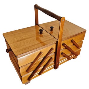 Pu&ograve; includere: Un portacucito in legno con finitura lucida, caratterizzato da un design a fisarmonica a pi&ugrave; livelli con scomparti, due coperchi superiori con piccoli pomelli rotondi e una maniglia centrale ad arco per il trasporto.