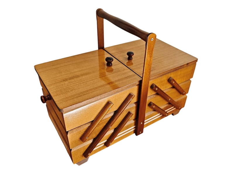 Pu&ograve; includere: Un portacucito in legno con finitura lucida, caratterizzato da un design a fisarmonica a pi&ugrave; livelli con scomparti, due coperchi superiori con piccoli pomelli rotondi e una maniglia centrale ad arco per il trasporto.