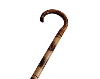 May include: A wooden walking cane featuring a curved handle and a cylindrical shaft. The wood displays a natural light tan colour with dark brown, scorched-looking grain patterns and horizontal black ring accents along the upper section.