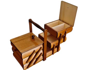 May include: A wooden sewing box with a cantilevered design featuring multiple tiered compartments. The box has a dark brown finish, a central carrying handle, and a hinged lid on the top section that opens to reveal a divided interior storage space.