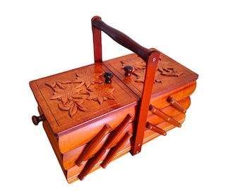 May include: A wooden sewing box with a cantilevered accordion design and a central carrying handle. The two top lids feature carved floral motifs and small round knobs. The box has multiple tiered compartments that expand outward when opened.