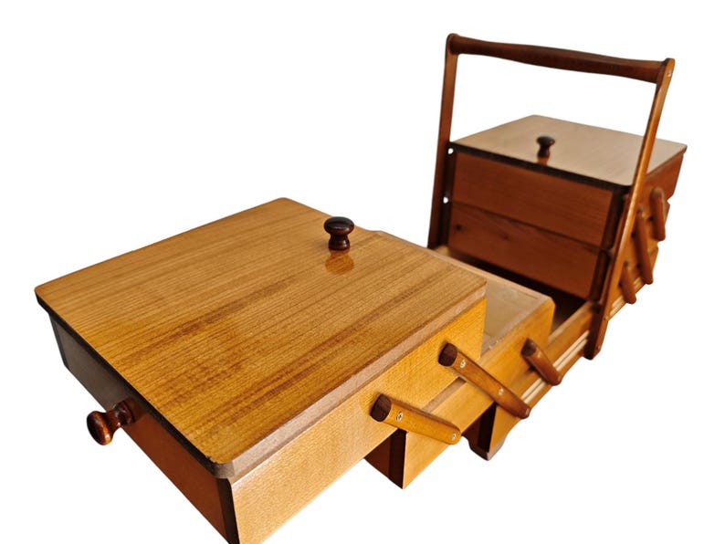 Pu&ograve; includere: Un portacucito in legno con un design a livelli estensibile dotato di scomparti multipli. La scatola presenta una finitura lucida color marrone chiaro, un manico alto e piccoli pomelli marrone scuro sui coperchi. &Egrave; in posizione aperta.