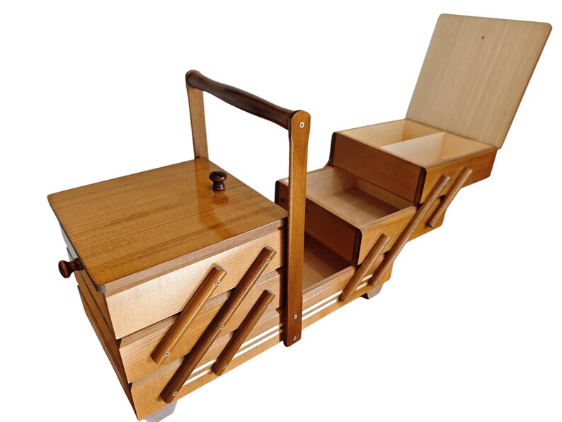 Pu&ograve; includere: Un portacucito in legno con un design a livelli estensibile dotato di molteplici scomparti. La scatola presenta una finitura lucida color marrone chiaro, una maniglia centrale per il trasporto e piccoli pomelli scuri sui coperchi.