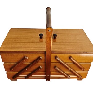 Pu&ograve; includere: Un portacucito rettangolare in legno con un design a livelli estensibile. La scatola presenta una finitura liscia color marrone chiaro, un manico verticale centrale e due piccoli pomelli scuri sul coperchio. I lati hanno bracci di supporto.