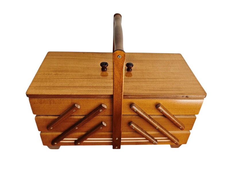 Pu&ograve; includere: Un portacucito rettangolare in legno con un design a livelli estensibile. La scatola presenta una finitura liscia color marrone chiaro, un manico verticale centrale e due piccoli pomelli scuri sul coperchio. I lati hanno bracci di supporto.