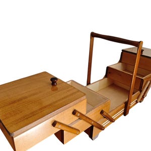 Pu&ograve; includere: Un portacucito in legno con finitura marrone chiaro, caratterizzato da un design a sbalzo con molteplici scomparti a livelli che si espandono verso l'esterno. La scatola include un'alta maniglia per il trasporto e pomelli marrone scuro.