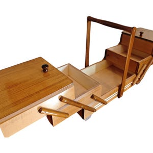 Pu&ograve; includere: Un portacucito in legno con design a soffietto, dotato di molteplici scomparti a livelli che si espandono verso l'esterno. La scatola presenta una finitura lucida color marrone chiaro, un'alta maniglia centrale e piccoli pomelli scuri sui coperchi.