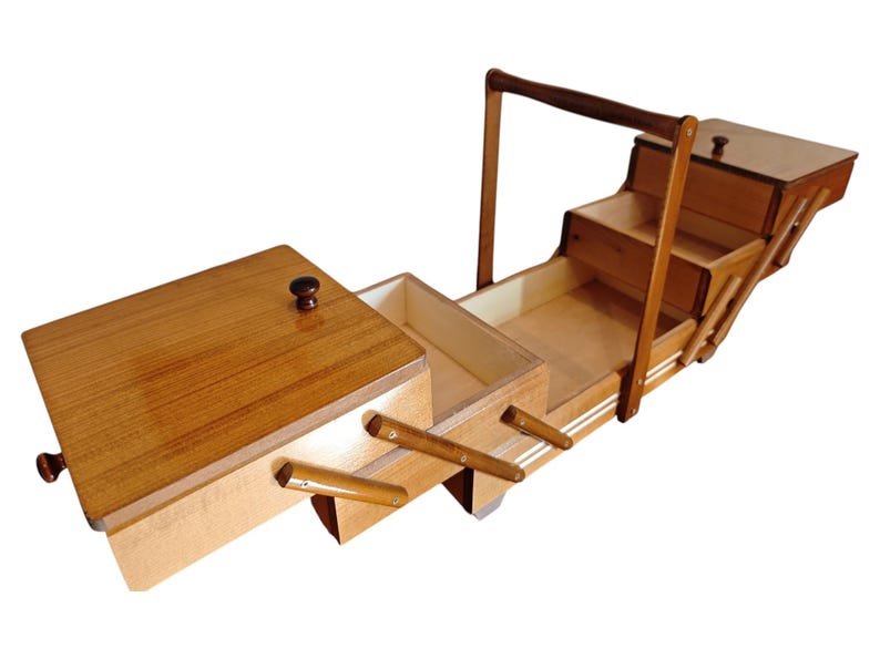 Pu&ograve; includere: Un portacucito in legno con design a soffietto, dotato di molteplici scomparti a livelli che si espandono verso l'esterno. La scatola presenta una finitura lucida color marrone chiaro, un'alta maniglia centrale e piccoli pomelli scuri sui coperchi.