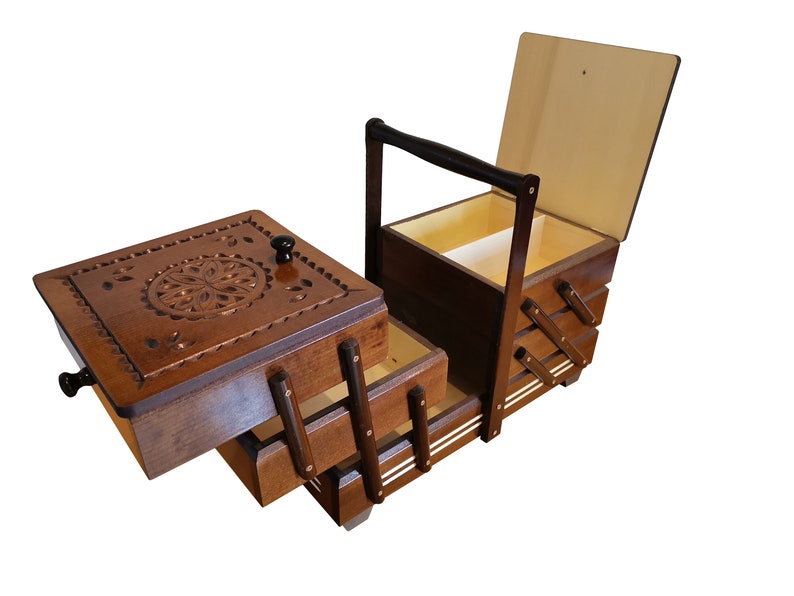 Puede incluir: Una caja de costura de madera con tapa abatible y tres cajones. La caja tiene un dise&ntilde;o tallado en la parte superior y un asa en el lateral.