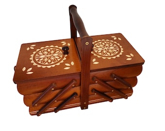 May include: A rectangular wooden sewing box with a tiered accordion-style design and a central carrying handle. The two top lids feature circular floral inlay patterns and small dark knobs, with additional leaf-shaped inlays on the surface.
