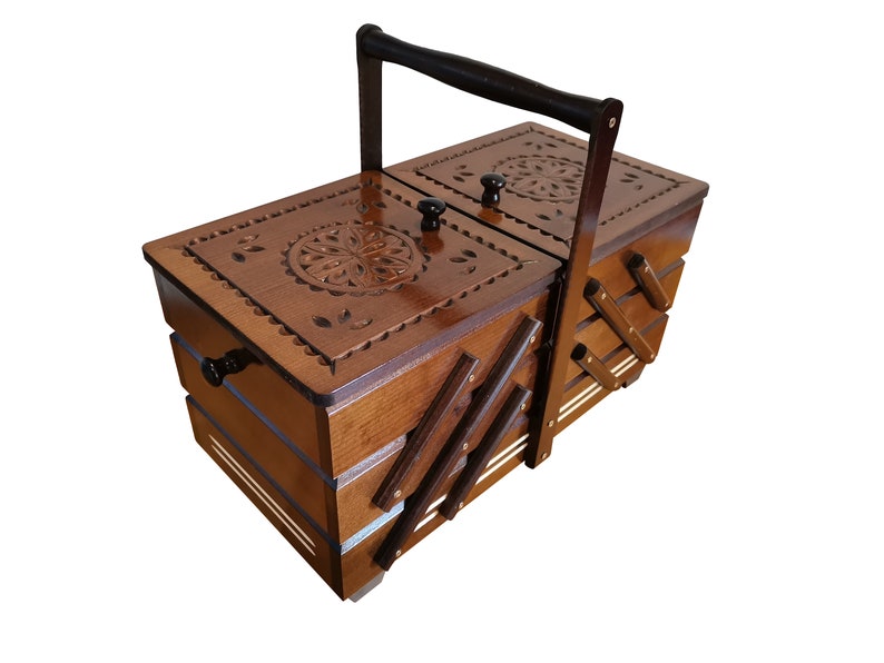 Puede incluir: Una caja de costura de madera con tapa abatible y asa de transporte. La caja tiene un dise&ntilde;o floral tallado en la parte superior y est&aacute; dividida en compartimentos.