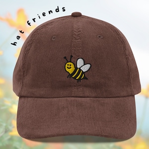 May include: Brown corduroy baseball cap with an embroidered yellow and black bee with a smiling face. The text "hat friends" is written in black above the bee.