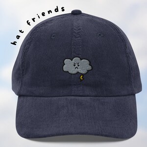 May include: A navy blue corduroy baseball cap with an embroidered gray cloud with a lightning bolt and a frowning face. The text "hat friends" is written in black on the side of the cap.