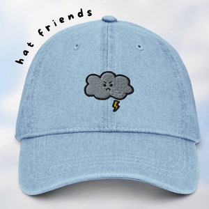 May include: A light blue denim baseball cap with an embroidered gray cloud with a yellow lightning bolt and a frowning face. The text "hat friends" is written in black on the side of the cap.