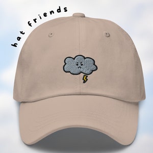 May include: A beige baseball cap with an embroidered gray cloud with a lightning bolt and a frowning face. The text "hat friends" is written in black above the cloud. The text "Art on a Bagel" is written in black on a white background in the lower right corner of the image.