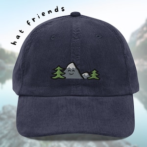 Puede incluir: Una gorra de béisbol de pana azul marino con un diseño bordado de una montaña sonriente con dos árboles verdes. El texto "hat friends" está escrito en el lateral de la gorra.