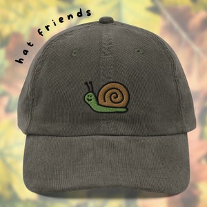 Puede incluir: Una gorra de béisbol de pana verde con un diseño de caracol bordado. El caracol es verde con una concha marrón y una cara sonriente. El texto "hat friends" está escrito en negro en el lateral de la gorra.