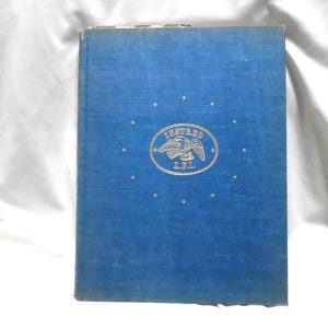 Può includere: Un libro con copertina rigida blu caratterizzato da un emblema centrale dorato di un'aquila all'interno di un ovale con il testo INSURED L.F.I. circondato da una disposizione circolare di piccole stelle dorate. La copertina ha una finitura in tessuto.