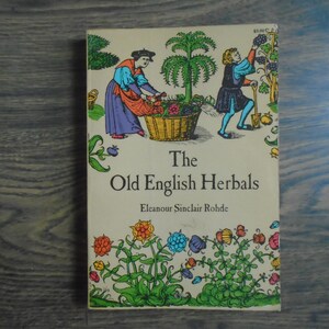 May include: A vintage book titled "The Old English Herbals" with illustrations of people harvesting plants. The cover features colorful drawings of flowers and herbs, with the author's name below the title. The book is set against a wooden background.