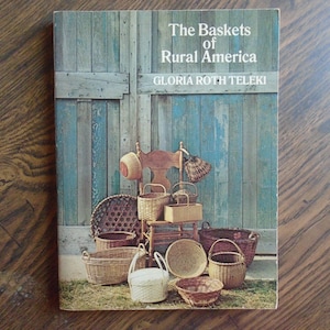 Puede incluir: Un libro titulado "The Baskets of Rural America" de Gloria Roth Teleki. La portada presenta una puerta de madera rústica con un acabado de pintura azul y una colección de cestas tejidas dispuestas en una silla de madera.