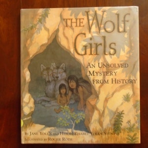 May include: Book cover with an illustration of two girls and two wolves in a cave. The title is "The Wolf Girls: An Unsolved Mystery From History" by Jane Yolen and Heidi Elisabet Yolen Stemple, illustrated by Roger Roth.