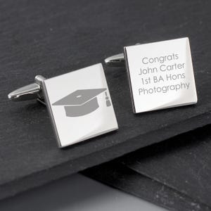 Könnte beinhalten: Quadratische Manschettenknöpfe aus Silber. Einer zeigt eine Grafik einer Graduiertenkappe, der andere den Text "Congrats John Carter 1st BA Hons Photography". Die Manschettenknöpfe liegen auf einer dunkelgrauen Oberfläche.