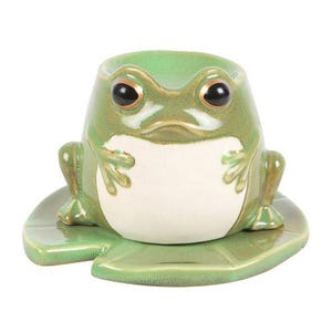 Könnte beinhalten: Ein keramikfroschförmiger Pflanztopf in Grün- und Weißtönen. Der Frosch hat große, dunkle Augen und sitzt auf einer Seerosenblattbasis. Der Pflanztopf ist mit einem hohlen Innenraum zur Aufnahme von Pflanzen oder anderen Dekorationsgegenständen versehen.