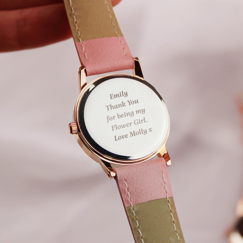 May include: A rose gold wristwatch with a white face and a pink and tan leather strap. The watch face has the inscription "Emily Thank You for being my Flower Girl. Love Molly x."