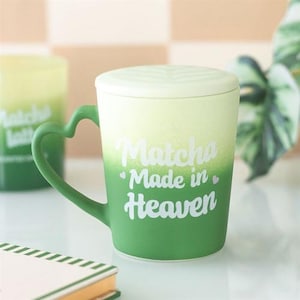 Puede incluir: Una taza de cerámica degradada verde y blanca con un asa en forma de corazón y una tapa a juego. La taza presenta el texto "Matcha Made in Heaven" en escritura blanca. La taza está diseñada para bebidas.