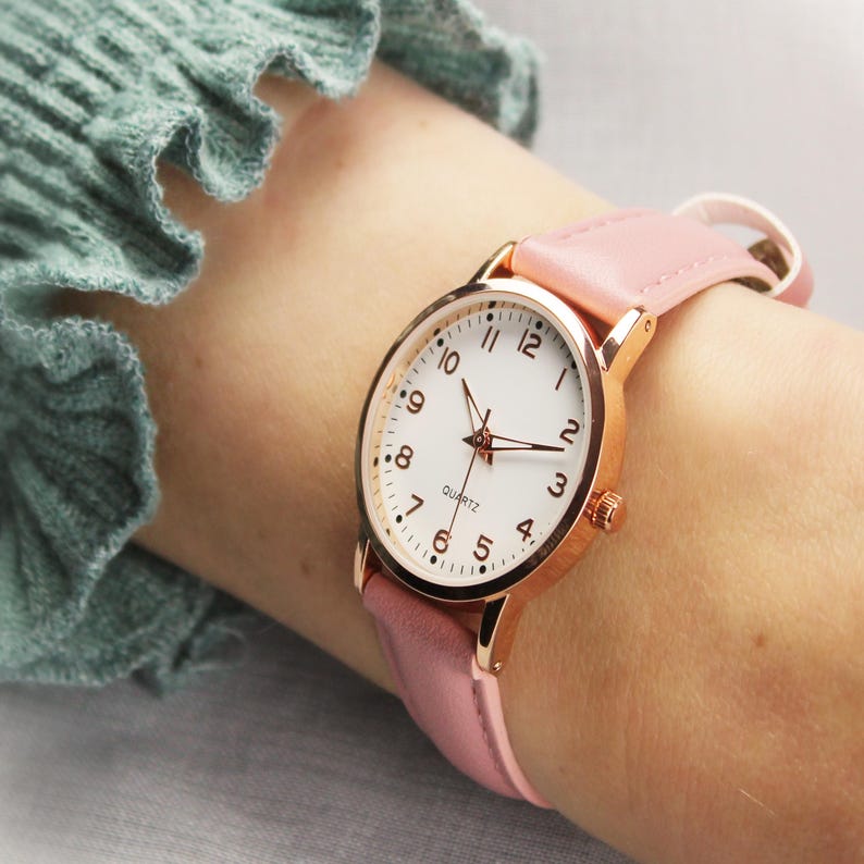 May include: A rose gold quartz watch with a white face and black numerals. The watch has a pink leather band. The watch is on a wrist.
