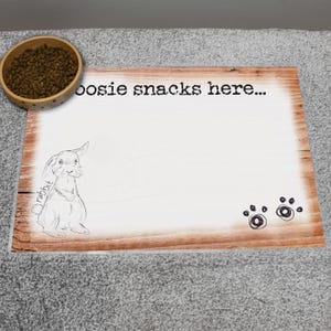 May include: A wooden pet food mat with the text "boosie snacks here..." and a sketch of a rabbit. The mat has a light wood grain pattern and includes two paw prints. A wooden bowl filled with pet food is in the upper left corner.