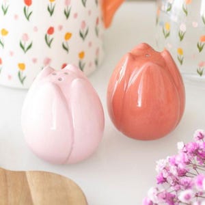 Op de afbeelding: Twee zout- en peperstrooiers in de vorm van tulpen. Eén is lichtroze, de andere koraal. Beide hebben drie gaten om te doseren. Op de achtergrond staan keramische items met bloemmotieven. Ze staan op een wit oppervlak.