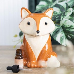 May include: A ceramic fox-shaped essential oil diffuser in orange and cream. The fox has black eyes and a white chest and tail. A small brown bottle with a dropper is next to the diffuser. The bottle has a label with text.