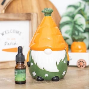 Op de afbeelding: Een keramische dwerg-vormige etherische olie diffuser met een oranje wortelvormige hoed en een groene body versierd met witte bloemen. Een klein bruin flesje etherische olie met een druppelaar staat naast de diffuser.